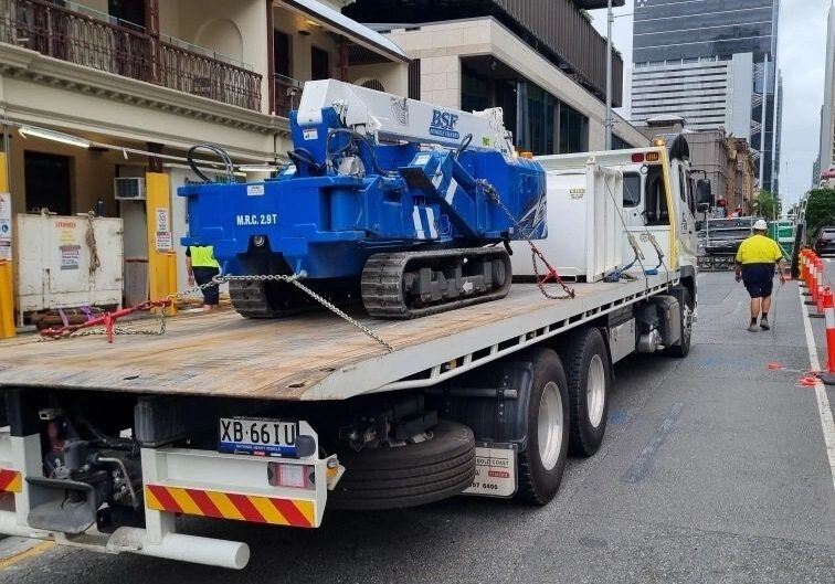 Truck transporting blue construction equipment in city.
